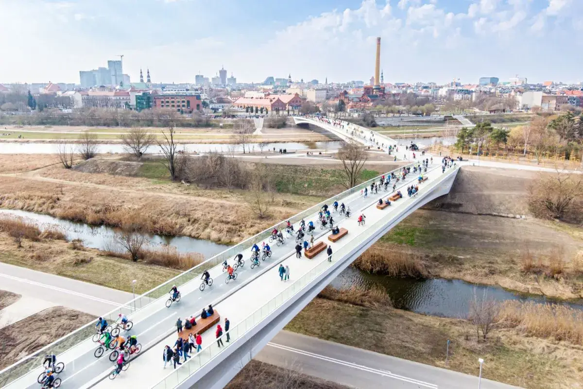 Rowerzyści i spacerowicze na nowoczesnym moście w Poznaniu, z panoramą miasta w tle.