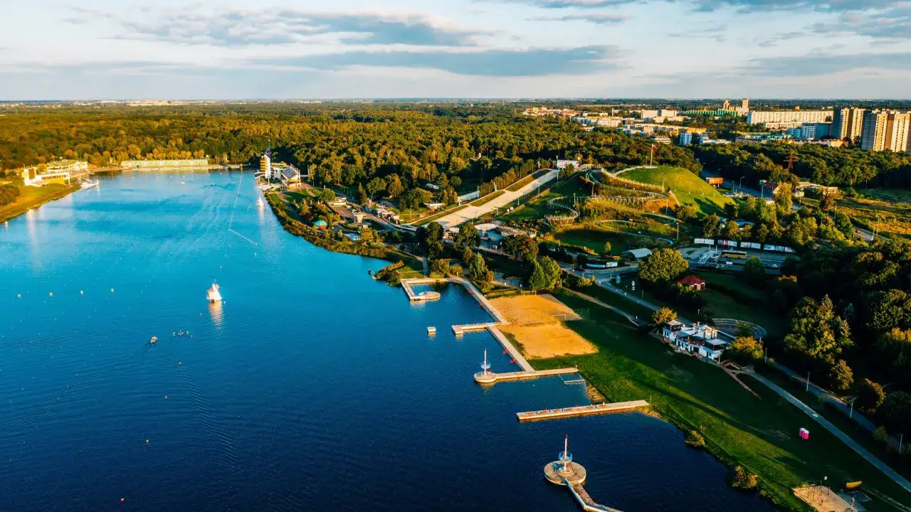 Malownicze jezioro do pływania otoczone lasem, z piaszczystą plażą, pomostami i budynkami rekreacyjnymi. W oddali widać miasto.