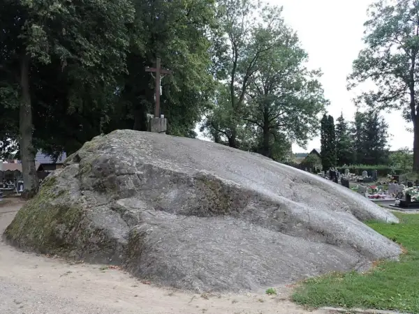 Ogromny głaz narzutowy na cmentarzu, zwieńczony krzyżem. Otoczony drzewami i nagrobkami.